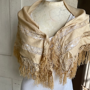 May include: A cream-colored lace shawl with fringe trim. The shawl is made of a lightweight fabric and has a delicate lace pattern.