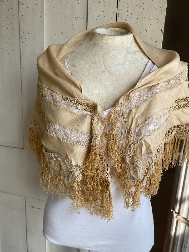 May include: A cream-colored lace shawl with fringe trim. The shawl is made of a lightweight fabric and has a delicate lace pattern.