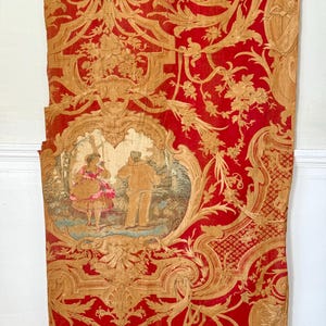 May include: A vintage red and gold fabric with a repeating pattern of stylized floral and scroll designs. A central oval features a scene of two figures in a landscape.