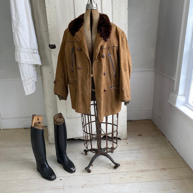 May include: A brown leather jacket with a fur collar on a vintage clothing display stand. The jacket has a double breasted closure and two pockets on the front. The stand is made of metal and has a round base.