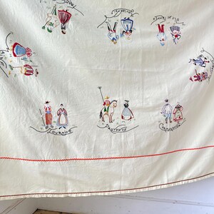 May include: A white linen tablecloth with red embroidered borders and colorful embroidered figures representing different regions of France. The regions are labeled in French: "Bretagne", "Lorraine", "Provence", "Champagne", "Alsace", "Normandie", and "Savoie".