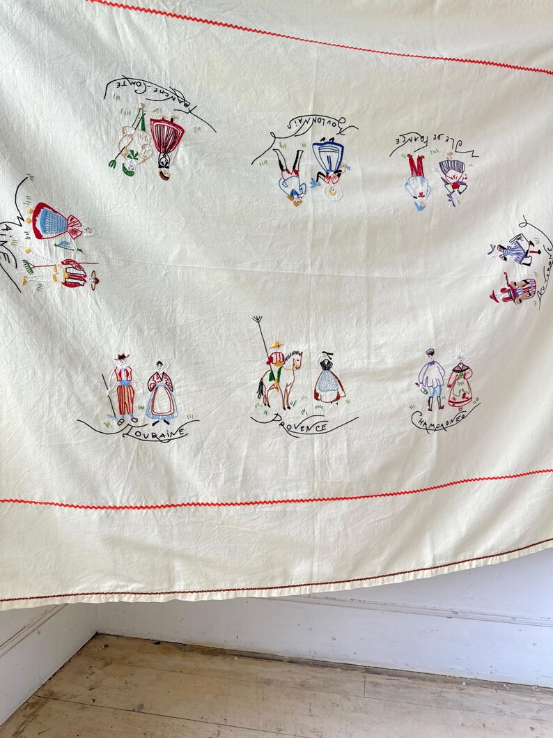 May include: A white linen tablecloth with red embroidered borders and colorful embroidered figures representing different regions of France. The regions are labeled in French: "Bretagne", "Lorraine", "Provence", "Champagne", "Alsace", "Normandie", and "Savoie".