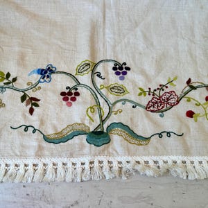 May include: A white linen tablecloth with embroidered floral designs in shades of green, red, purple, and blue. The tablecloth has a white fringe border.