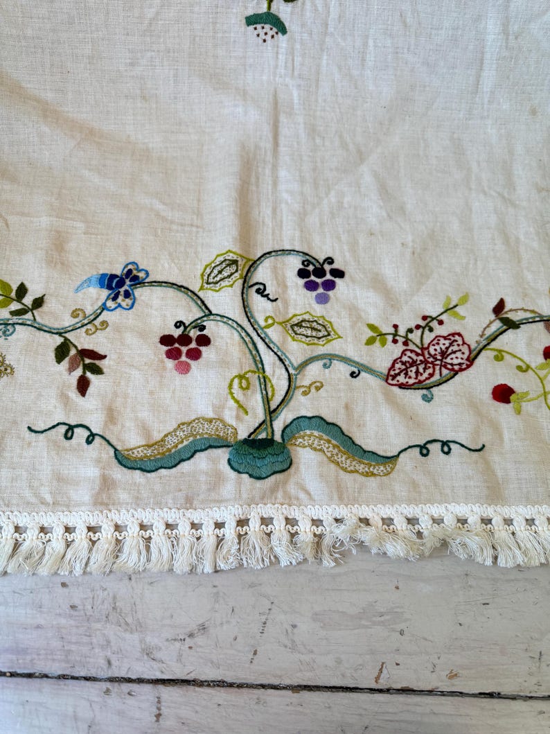 May include: A white linen tablecloth with embroidered floral designs in shades of green, red, purple, and blue. The tablecloth has a white fringe border.