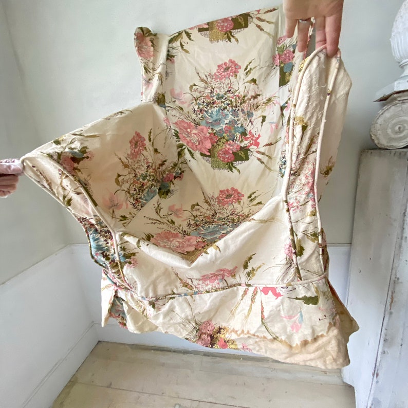 May include: A vintage floral fabric with a cream background and pink, blue, and green flowers. The fabric has a ruffled edge and is folded in half.