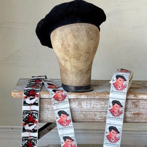 Peut inclure: Un béret en laine noir est posé sur une tête de mannequin. Des rouleaux de ruban avec l'image d'un homme portant un béret et le nom "Juantzito" sont sur une surface en bois.