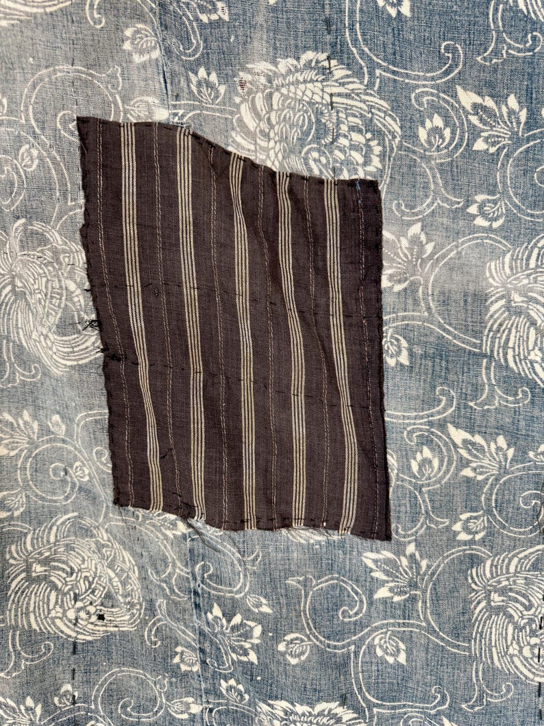 May include: A close-up of a blue and white fabric with a brown and white striped patch sewn on. The fabric has a floral pattern with a repeating design of stylized flowers and leaves.