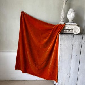 Op de afbeelding: Een oranje fluwelen stof gedrapeerd over een witte, verweerde kast. Een decoratieve witte pilaar en een gebeeldhouwde bekroning staan bovenop de kast. De stof heeft een rijke, warme kleur en een zachte textuur.