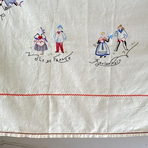 May include: White linen tablecloth with red embroidered trim and colorful embroidered figures of people in traditional French clothing. The text "Ile de France" and "Boulognais" is embroidered on the tablecloth.