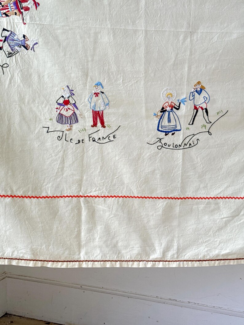 May include: White linen tablecloth with red embroidered trim and colorful embroidered figures of people in traditional French clothing. The text "Ile de France" and "Boulognais" is embroidered on the tablecloth.