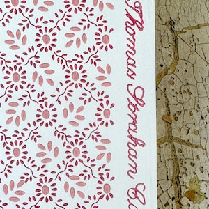 Può includere: Sezione di carta bianca con un motivo ripetuto di fiori e foglie rosa. Le parole "Thomas Graham Co." sono scritte in corsivo nello stesso colore rosa. Il lato destro dell'immagine mostra una superficie strutturata marrone chiaro.