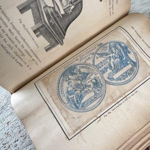 Pode incluir: Um close-up de uma página de um livro antigo com uma ilustração em preto e branco de uma máquina. A página também apresenta dois selos circulares azuis e brancos com o texto "Exposition Universelle 1889" e "Republique Francaise" em francês.