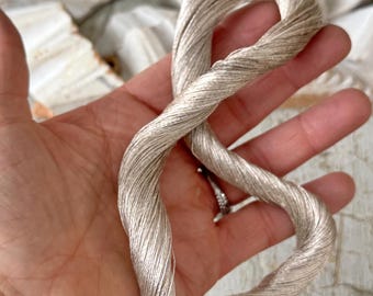 May include: A skein of light silver-gray thread, tightly twisted, is held in a hand. The thread has a subtle sheen, suggesting a silk or similar fiber. The end of the thread shows a small red and white tassel. The background is blurred.