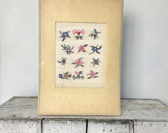 May include: A framed artwork featuring a collection of 16 embroidered floral designs in shades of pink, blue, and green. The artwork is displayed on a white wooden surface.
