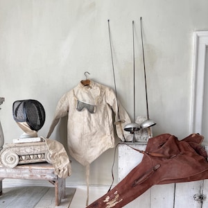 Puede incluir: Colección de equipo de esgrima antiguo, que incluye una chaqueta color crema, una máscara de malla negra, guantes y tres espadas. Una funda de espada de cuero marrón descansa sobre un mueble blanco. Los artículos están colocados contra una pared gris claro.