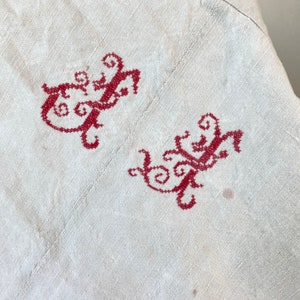 May include: A white linen fabric with two red embroidered designs. The designs are intricate and feature swirls and loops.