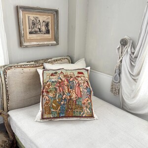 May include: A decorative pillow with a medieval tapestry design featuring figures in historical clothing. The pillow is placed on a white linen-covered daybed. A framed print hangs on the wall above the bed, and a curtain is draped to the right.