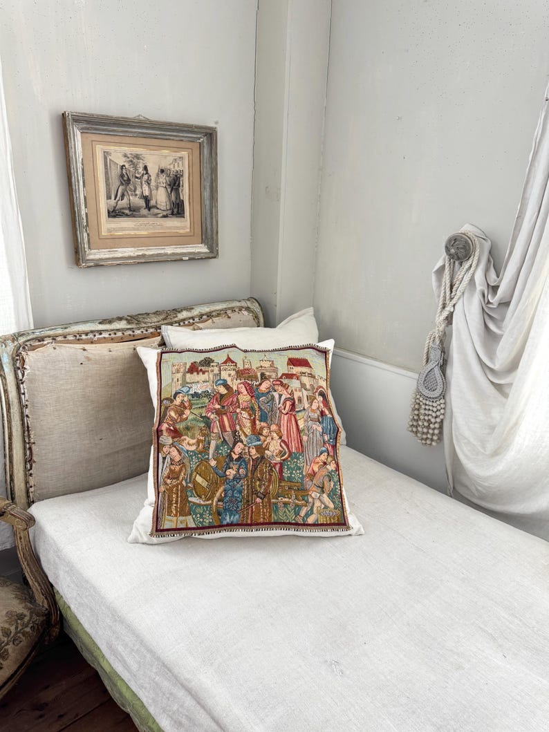 May include: A decorative pillow with a medieval tapestry design featuring figures in historical clothing. The pillow is placed on a white linen-covered daybed. A framed print hangs on the wall above the bed, and a curtain is draped to the right.
