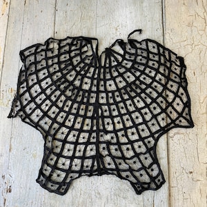 May include: Black lace shawl with a spiderweb design. The shawl is made of delicate black lace and has a circular shape.