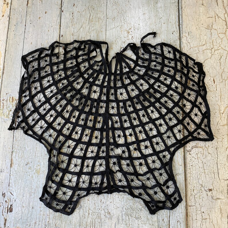 May include: Black lace shawl with a spiderweb design. The shawl is made of delicate black lace and has a circular shape.