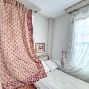 Może przedstawiać: Białe łóżko z białą pościelą lnianą i czerwoną i kremową wzorzystą zasłoną w tle. Łóżko znajduje się w pokoju z białymi ścianami i oknem z białymi zasłonami.