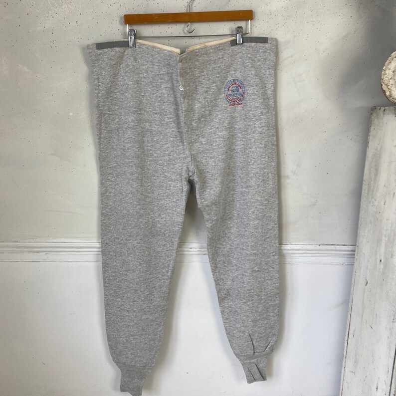 Pode incluir: Cal&ccedil;as de moletom cinzas com um bot&atilde;o de mosca e um pequeno logotipo bordado na perna esquerda. O logotipo &eacute; vermelho, branco e azul e diz "The Champion Athletic Wear, Inc.  Rochester, NY."