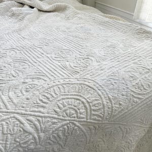 Peut inclure: Une courtepointe blanche et texturée avec des motifs floraux et géométriques complexes. La courtepointe est drapée sur un lit, avec un oreiller rayé visible en arrière-plan. Le design général est de style classique et élégant.
