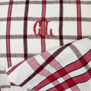 May include: A white fabric with red and black plaid pattern. The fabric has a monogram of the letters "CU" embroidered in red.