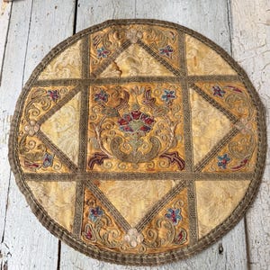 Puede incluir: Textil antiguo bordado, amarillo y marrón, de forma circular. El centro presenta un diseño floral ornamentado con detalles en rojo, azul y verde. El textil está dividido en secciones con un borde decorativo.