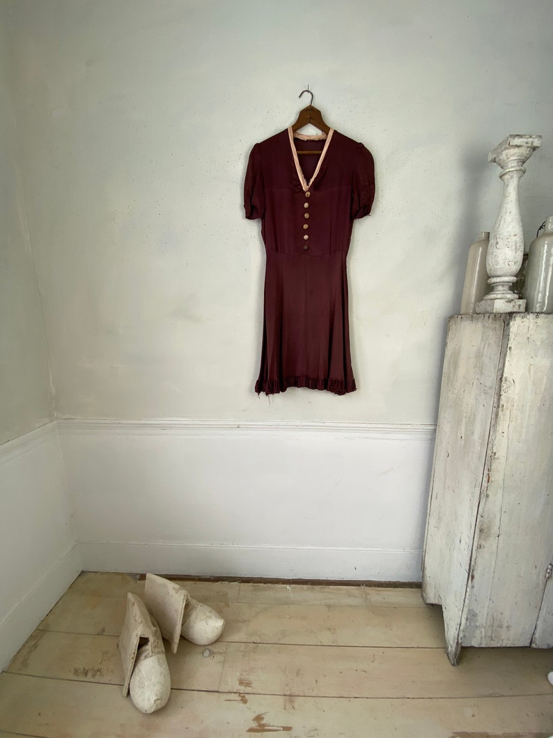 May include: A maroon vintage dress with a white collar and button fastening. The dress has short sleeves and a frilled hem. The dress is hanging on a wooden hanger.