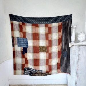 May include: A large patchwork quilt with a red and white plaid pattern. The quilt is made up of different fabrics, including a black and white striped fabric, a blue and white fabric, and a brown and white fabric. The quilt is hanging on a white wall.