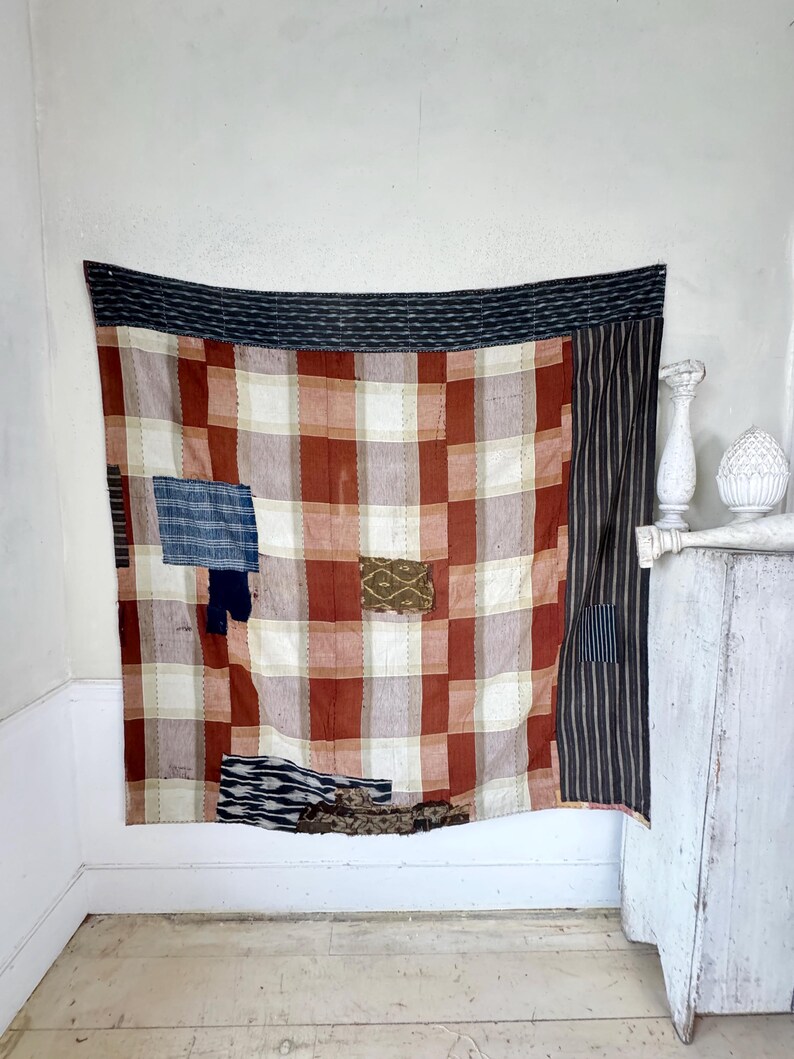May include: A large patchwork quilt with a red and white plaid pattern. The quilt is made up of different fabrics, including a black and white striped fabric, a blue and white fabric, and a brown and white fabric. The quilt is hanging on a white wall.