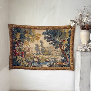 May include: A large tapestry depicting a landscape scene with a river, trees, and a building. The tapestry has a gold border and is mounted on a white wall. The scene includes birds and flowers, with a color palette of greens, blues, and beige.