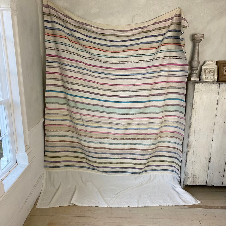 May include: A white striped throw blanket with a variety of colors, including pink, blue, and green. The blanket has a white fringe border and is draped over a white cabinet.
