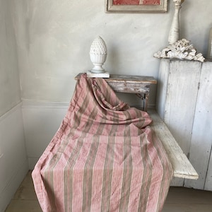 Può includere: Una tovaglia di tessuto a righe beige, rosso e bianco con un motivo a righe verticali. La tovaglia è drappeggiata su un tavolo di legno con una finitura in pino bianco.