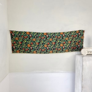 May include: A black tapestry with a floral pattern in shades of orange, red, green, and yellow. The tapestry is hanging on a white wall.