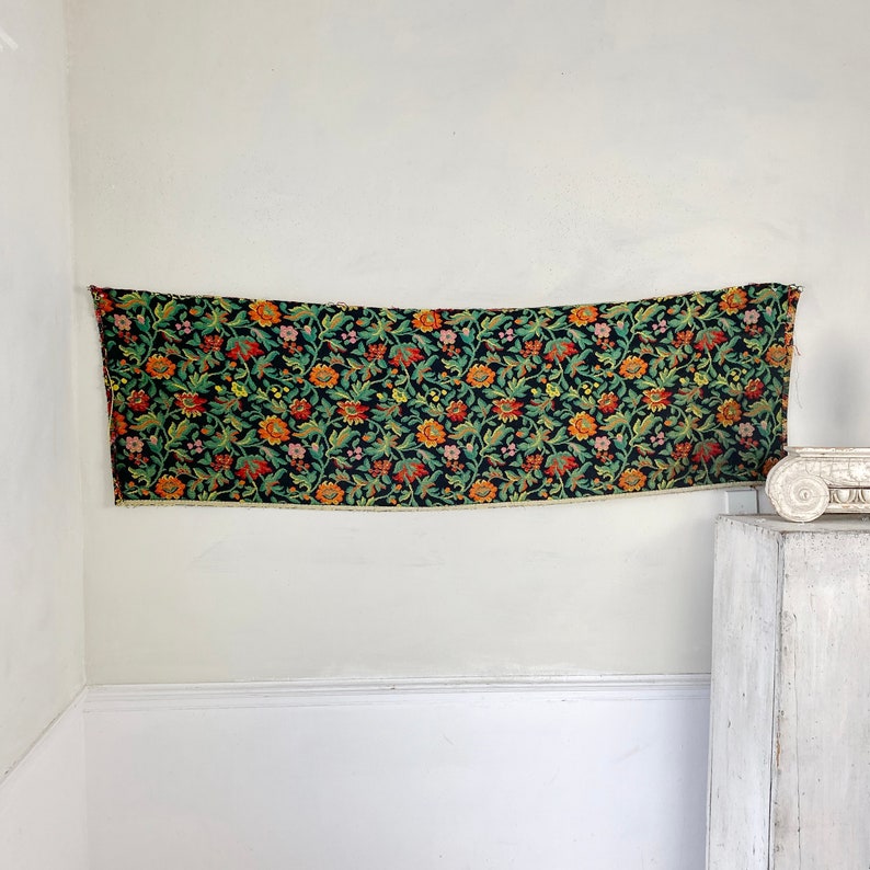 May include: A black tapestry with a floral pattern in shades of orange, red, green, and yellow. The tapestry is hanging on a white wall.