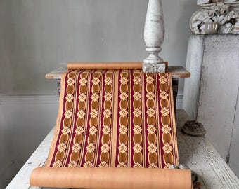 May include: A roll of vintage wallpaper with a repeating floral pattern in burgundy, gold, and tan. The wallpaper is displayed on a rustic wooden table, with a decorative white pillar in the background.