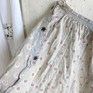 K&ouml;nnte beinhalten: Eine hellblaue Sch&uuml;rze mit einem wei&szlig;en und roten Blumenmuster. Die Sch&uuml;rze hat einen gerafften Rock und einen Bindeverschluss. Die Sch&uuml;rze ist aus einem leichten Stoff gefertigt und hat einen Vintage-Look.