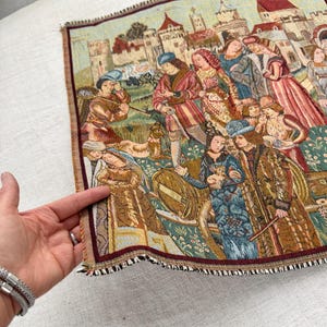 May include: A tapestry depicting a medieval scene with figures in period clothing, set against a backdrop of buildings and a landscape. The tapestry has a detailed, woven texture with a burgundy and brown fringe.