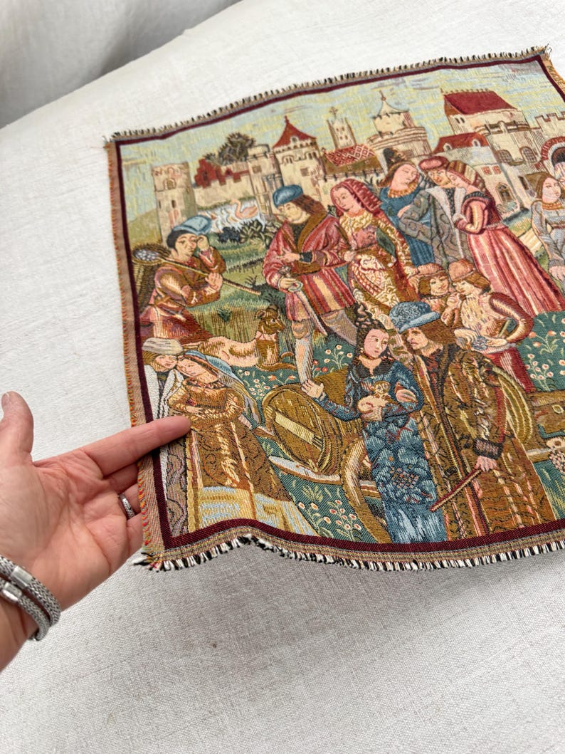 May include: A tapestry depicting a medieval scene with figures in period clothing, set against a backdrop of buildings and a landscape. The tapestry has a detailed, woven texture with a burgundy and brown fringe.