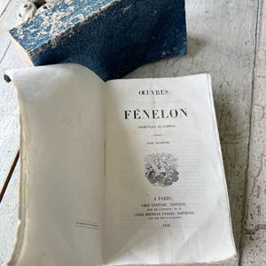 May include: An open antique book with the title "OEUVRES FÉNELON" printed in black ink. The book's cover is a distressed blue, and the pages are aged. The text "A PARIS" and the date "1838" are also visible.