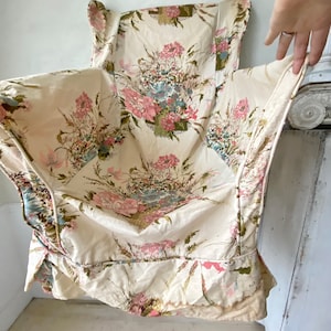 May include: A white fabric chair cushion with a floral print featuring pink, blue, and green flowers. The cushion has a ruffled edge and is made of a soft, textured fabric.