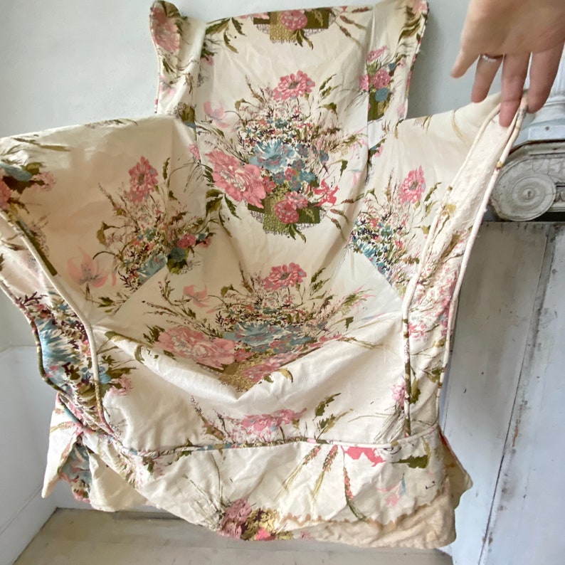 May include: A white fabric chair cushion with a floral print featuring pink, blue, and green flowers. The cushion has a ruffled edge and is made of a soft, textured fabric.