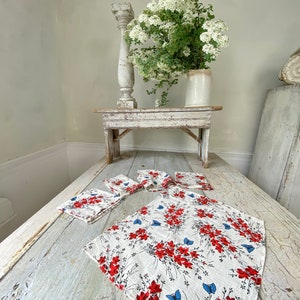 May include: A set of white linen napkins with a red floral and blue butterfly pattern. The napkins are folded and arranged on a whitewashed wooden table. A white vase with white flowers sits on a whitewashed wooden table in the background.