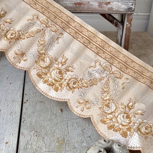 May include: Cream-colored vintage fabric with a scalloped edge, featuring a repeating pattern of roses, bows, and foliage in shades of brown and tan. The fabric has a decorative border and is likely intended for home decor.