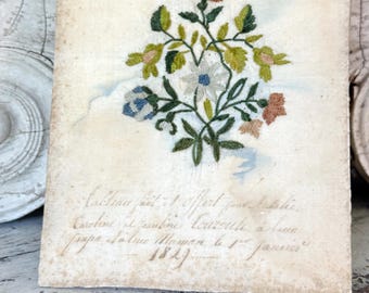 May include: Antique embroidered floral artwork on aged paper. The embroidery features a bouquet of flowers in shades of pink, blue, green, and white. The paper has handwritten text in French and the date 1827.