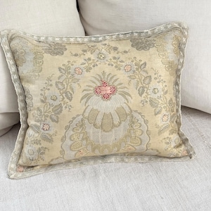 Puede incluir: Un cojín decorativo rectangular con un estampado floral en crema, beige y rosa pálido. El cojín tiene un diseño detallado con un borde festoneado y un motivo floral central. El cojín está colocado sobre una superficie blanca.