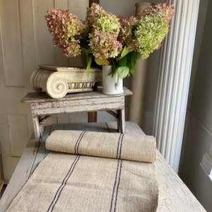 May include: A rustic, off-white linen runner with three dark blue stripes. The runner is rolled up on a white wooden table with a decorative white plaster column and a bouquet of pink and white hydrangeas.