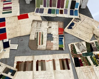 May include: A collection of fabric swatches in various colors and patterns, including red, blue, green, brown, and black. The swatches are arranged on a table with handwritten notes and labels.